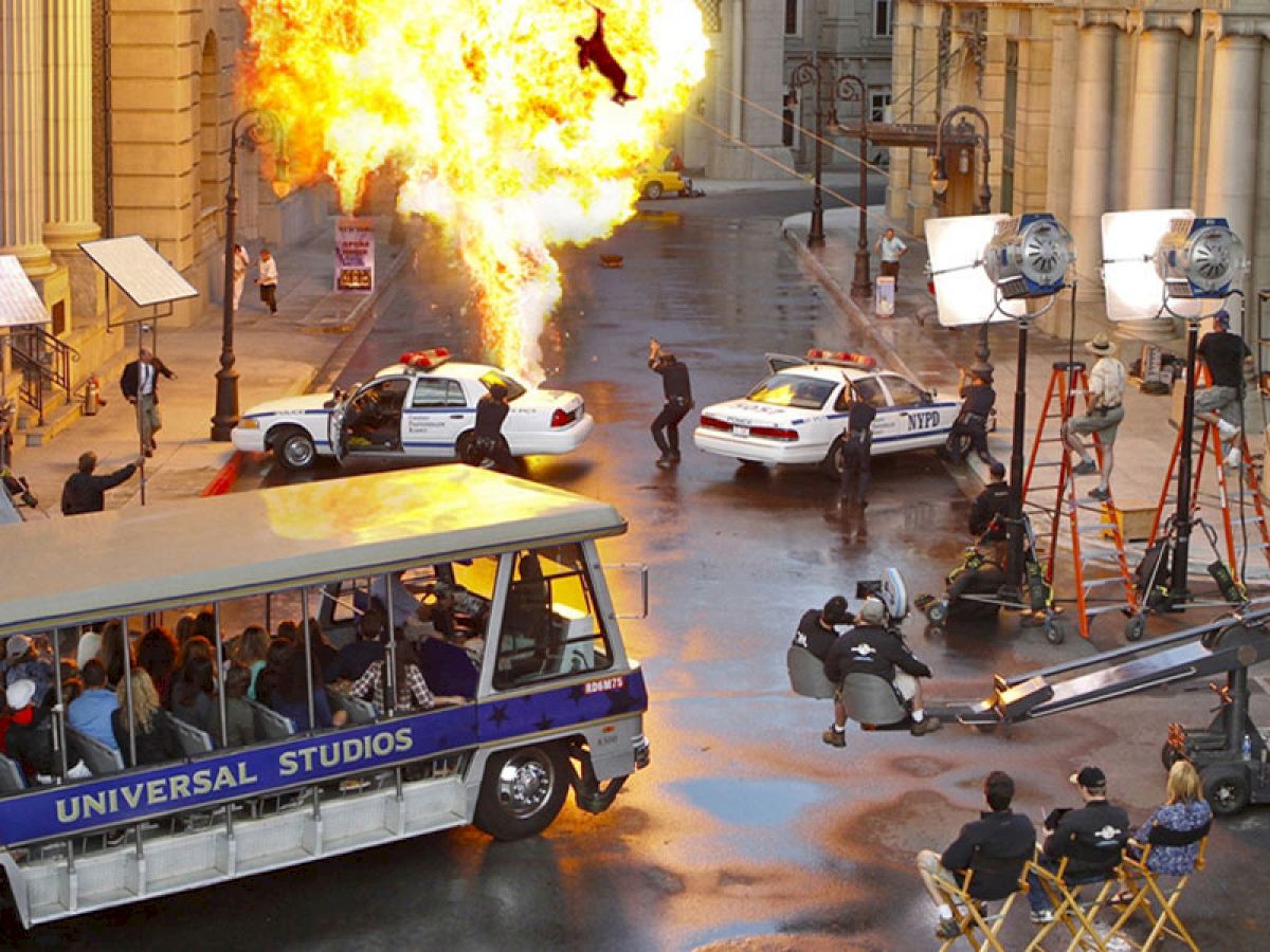 A film set featuring an explosion scene, police cars, a film crew, and a tram with 