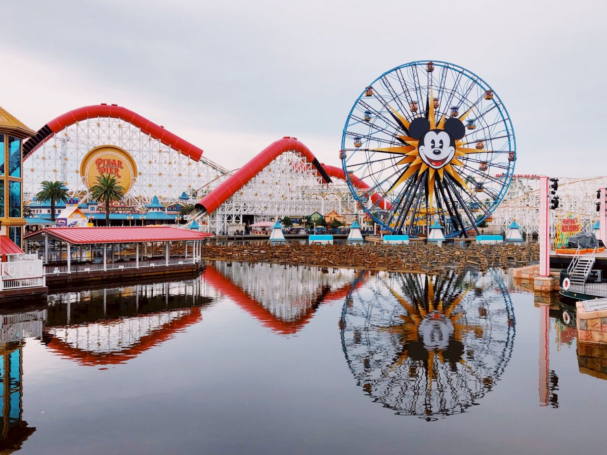 An amusement park with a Ferris wheel featuring a recognizable character's face, roller coasters, and a reflection in a calm body of water.