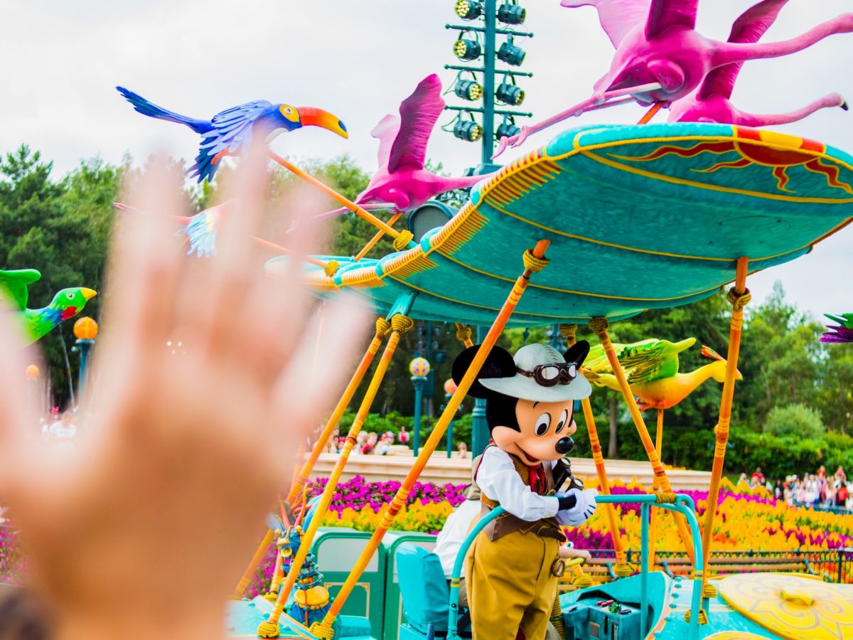 A theme park scene featuring a person in a mouse costume on a colorful float with birds, and a hand waving in the foreground.