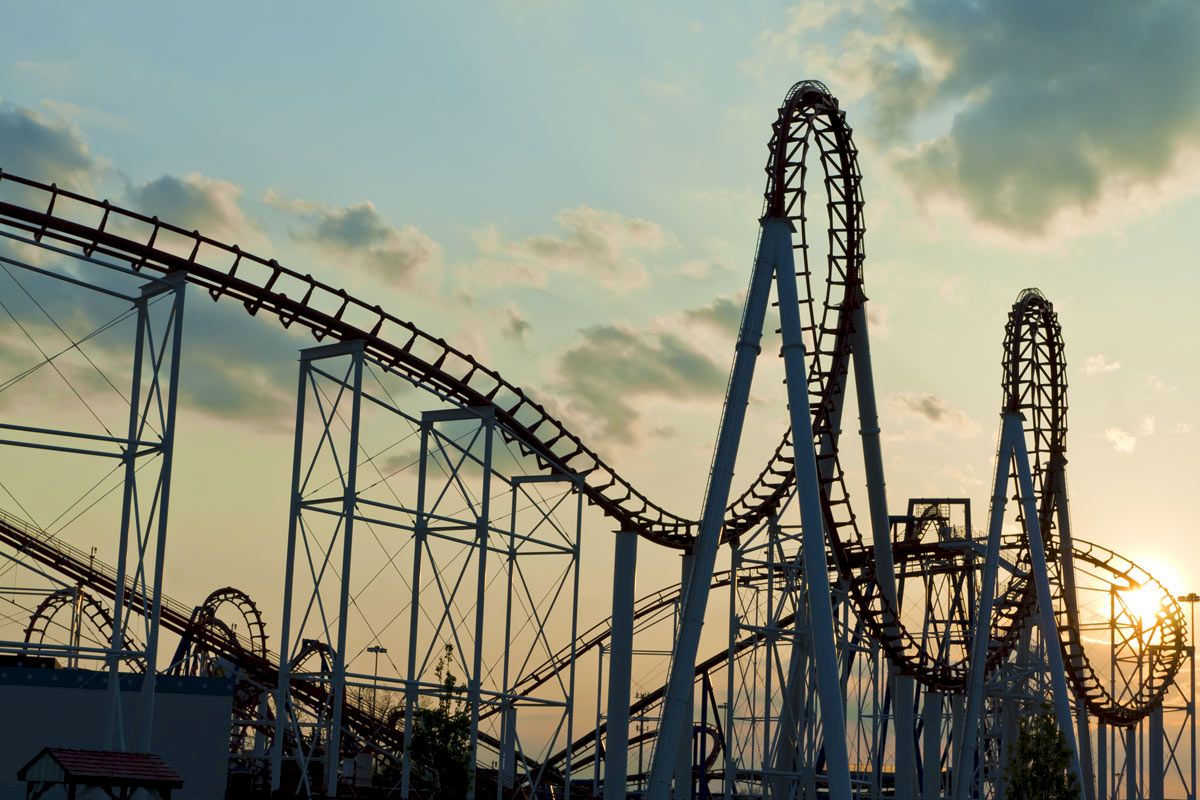 The image shows a roller coaster with multiple loops and tall steel structures silhouetted against a sunset sky with scattered clouds.