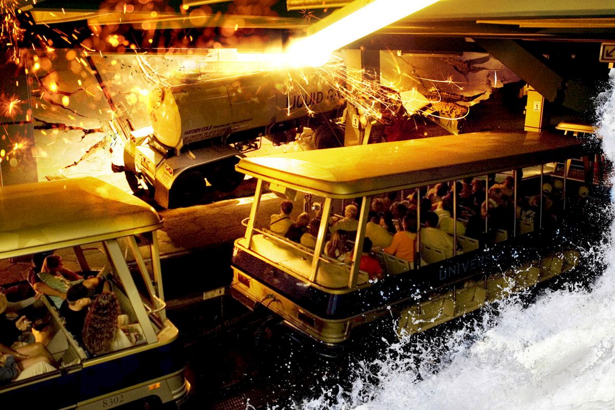 The image shows a theme park ride with passengers in a tram experiencing a simulated disaster scene involving fire, water, and destruction.