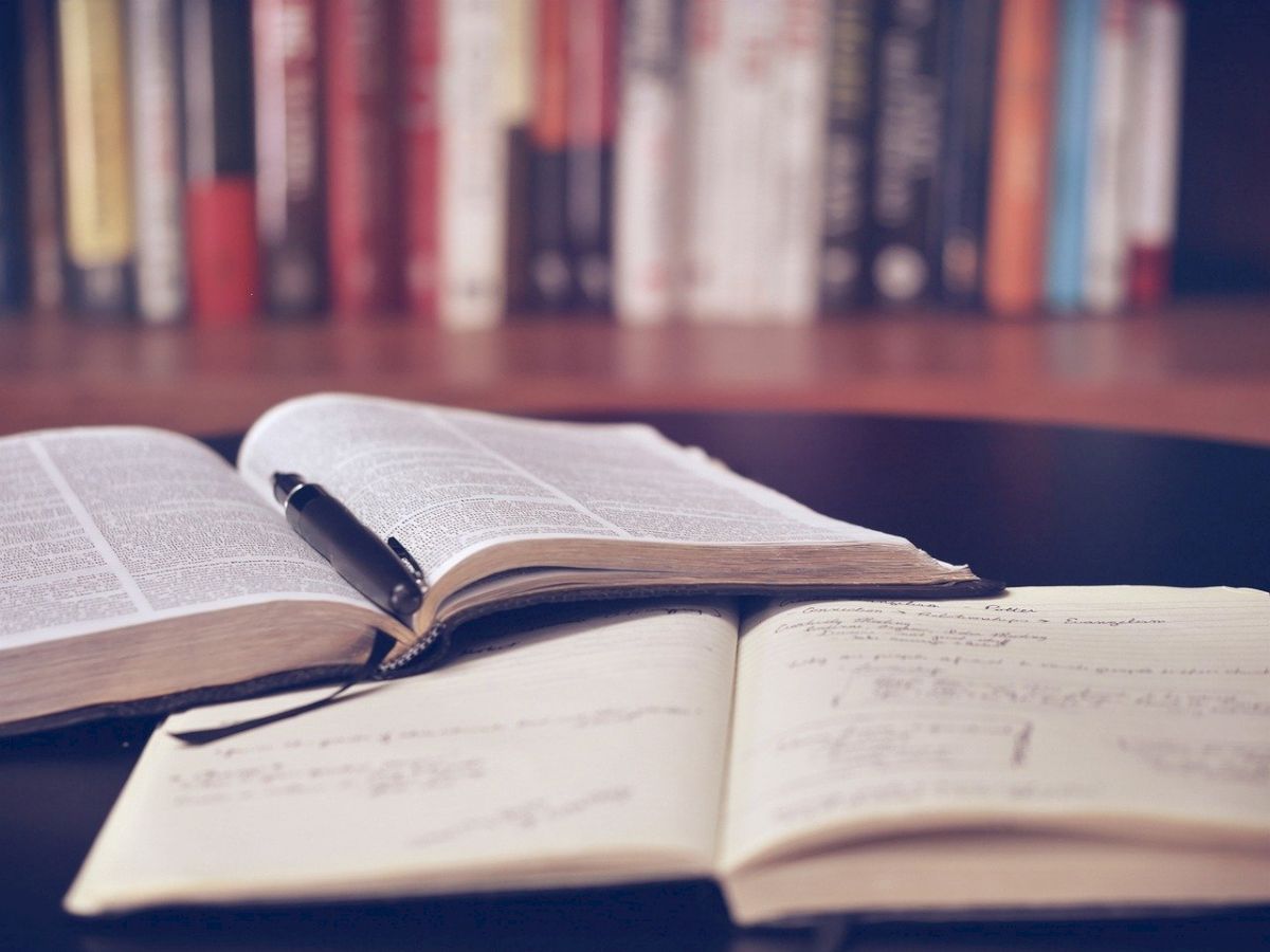 An open book with a pen on top and a notebook with writing are on a table, with a blurred background of books on a shelf.