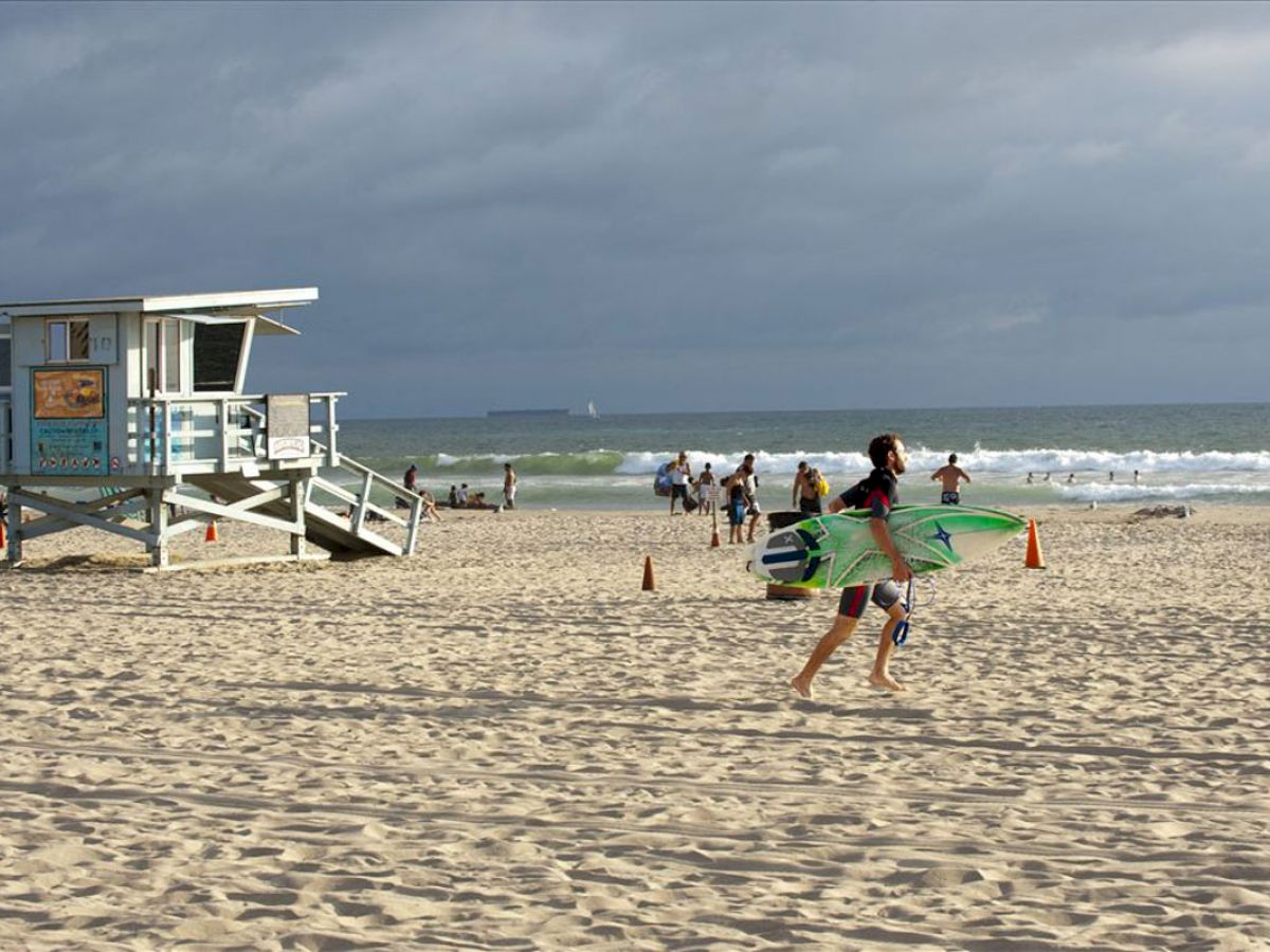 A lifeguard tower and people enjoying the beach, with one person carrying surfboards near the shoreline.