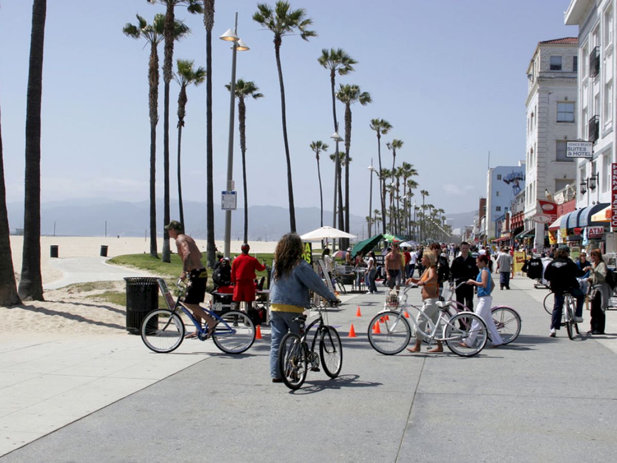 People riding bikes and strolling on a sunny boardwalk lined with palm trees, shops, and several vendors near the beach.