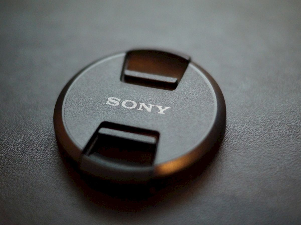 A Sony camera lens cap rests on a dark surface, displaying the brand name prominently in the center of the cap.