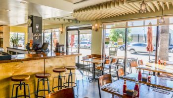 This image shows the interior of a modern, bright café or restaurant with wooden furniture, bar stools, and large windows allowing plenty of natural light.