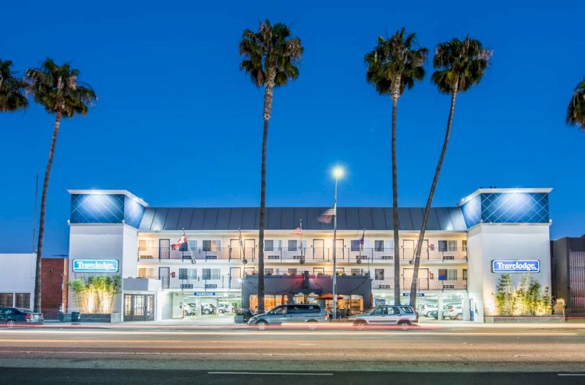 The image features a roadside hotel with prominent signage, surrounded by tall palm trees, and well-lit under a deep blue evening sky.