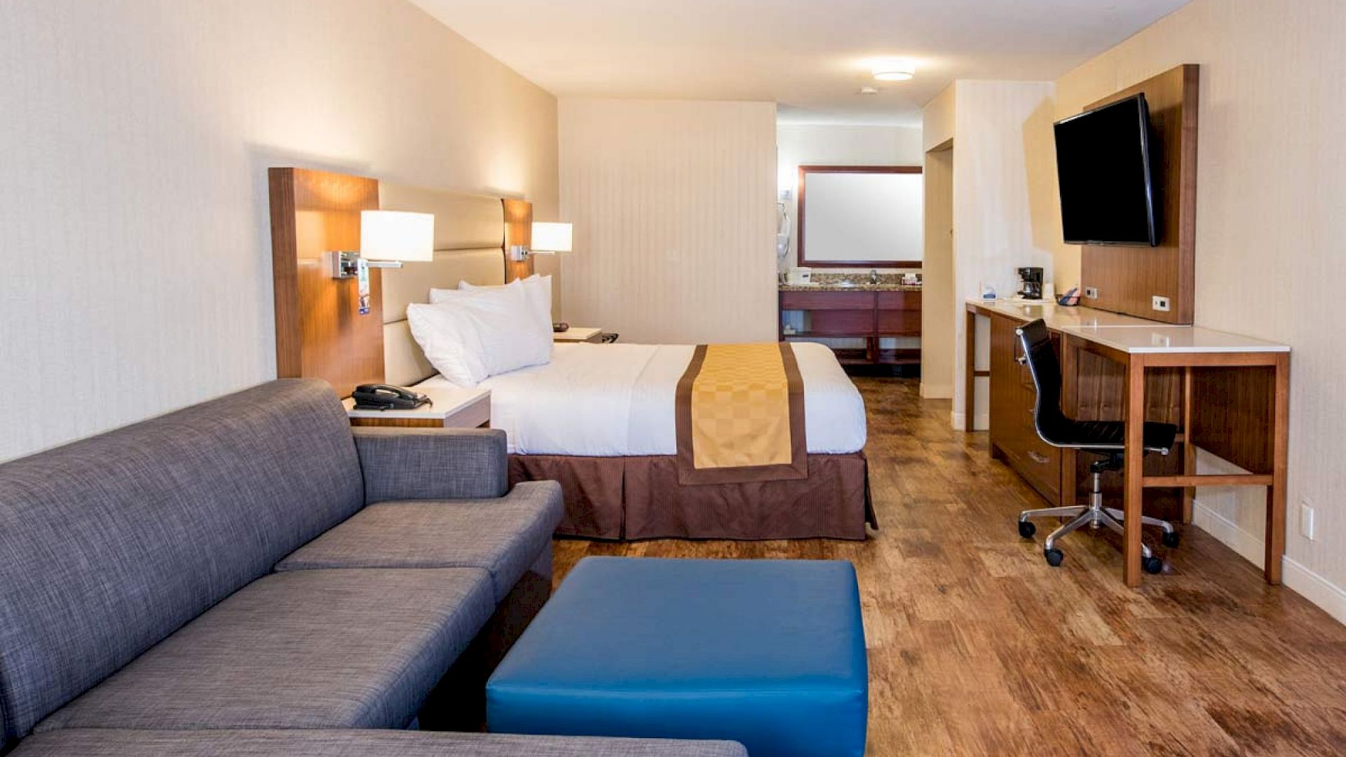 The image shows a modern hotel room with a bed, sofa, ottoman, desk, and wall-mounted TV. The room has wooden flooring and a clean design.