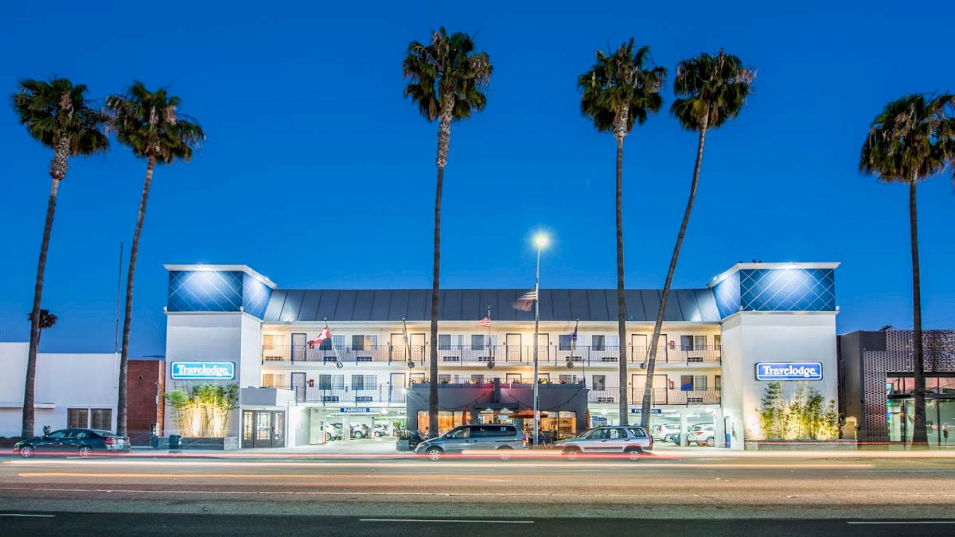 A multi-story hotel with palm trees in front is illuminated at night. Cars are parked at the entrance, and the sky is clear and dark blue.