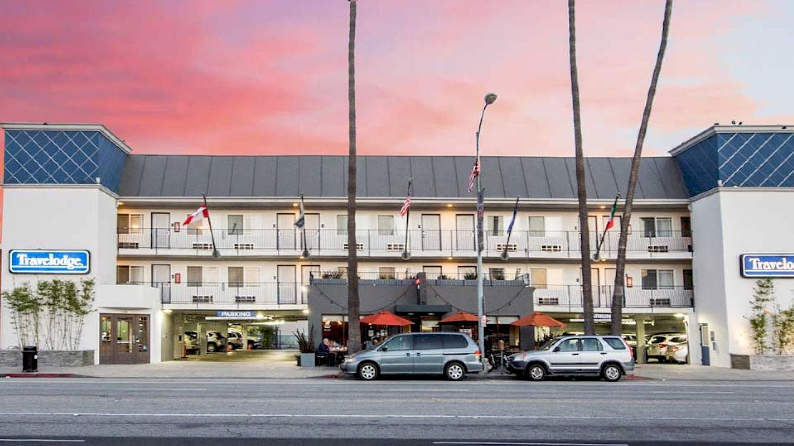 A Travelodge hotel with blue signs, palm trees, cars, and a pink sunset sky above the building.