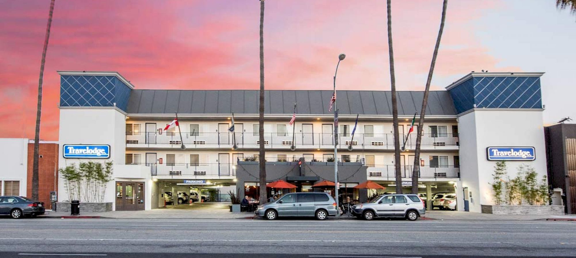 A Travelodge hotel with blue signs, palm trees, cars, and a pink sunset sky above the building.