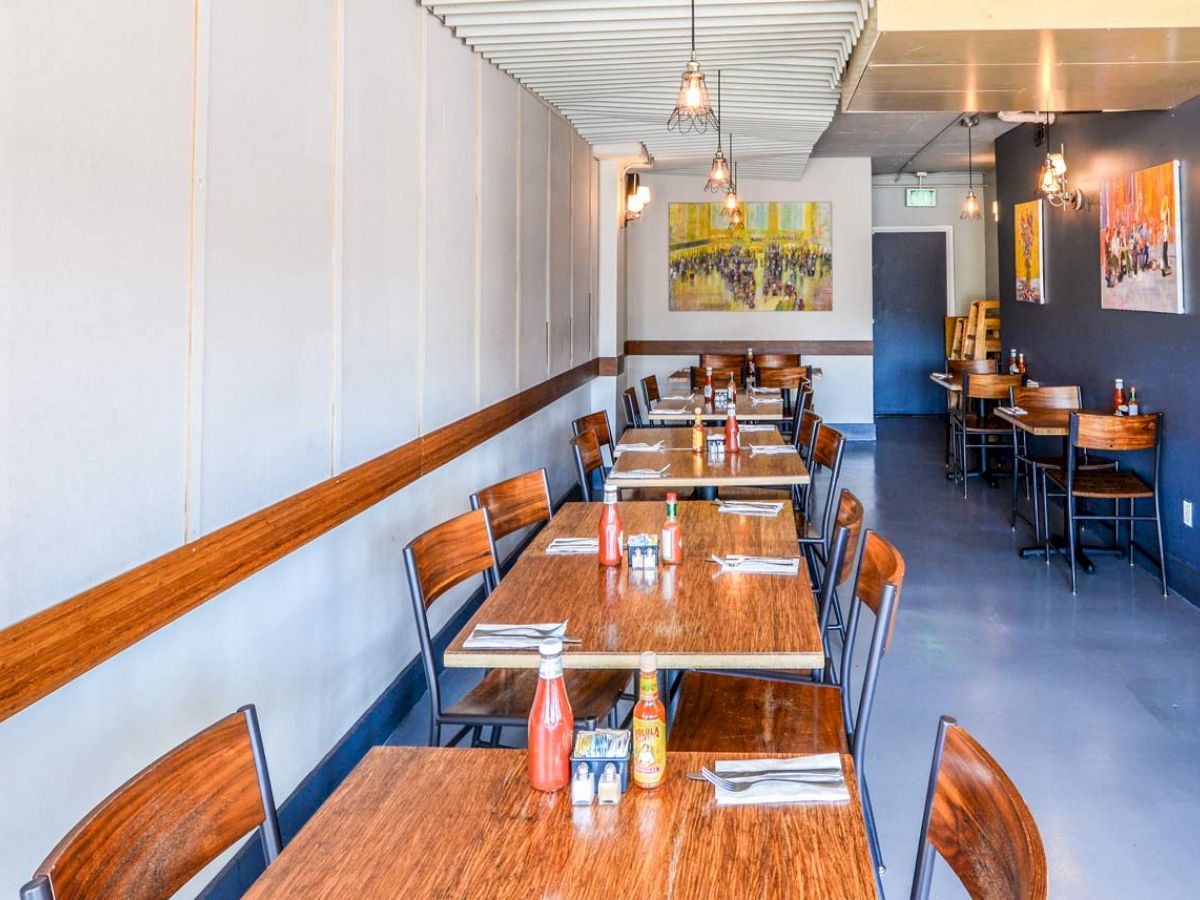 The image shows a modern restaurant with wooden tables and chairs, condiments on the tables, and artwork on the walls.