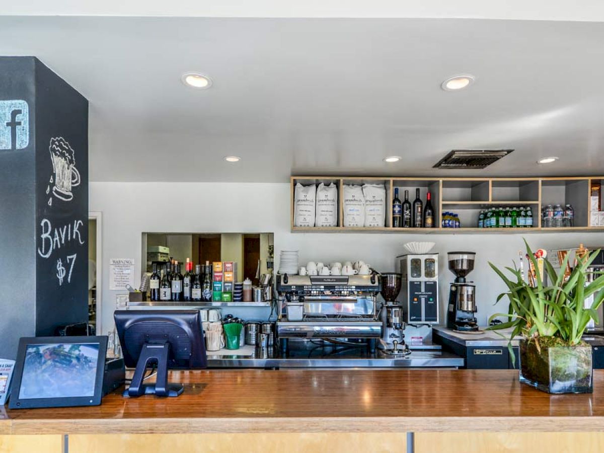 The image shows a coffee bar setup with various equipment, a cash register, and a tablet. Shelf with cups and bottles, some flyers, and plants are visible.