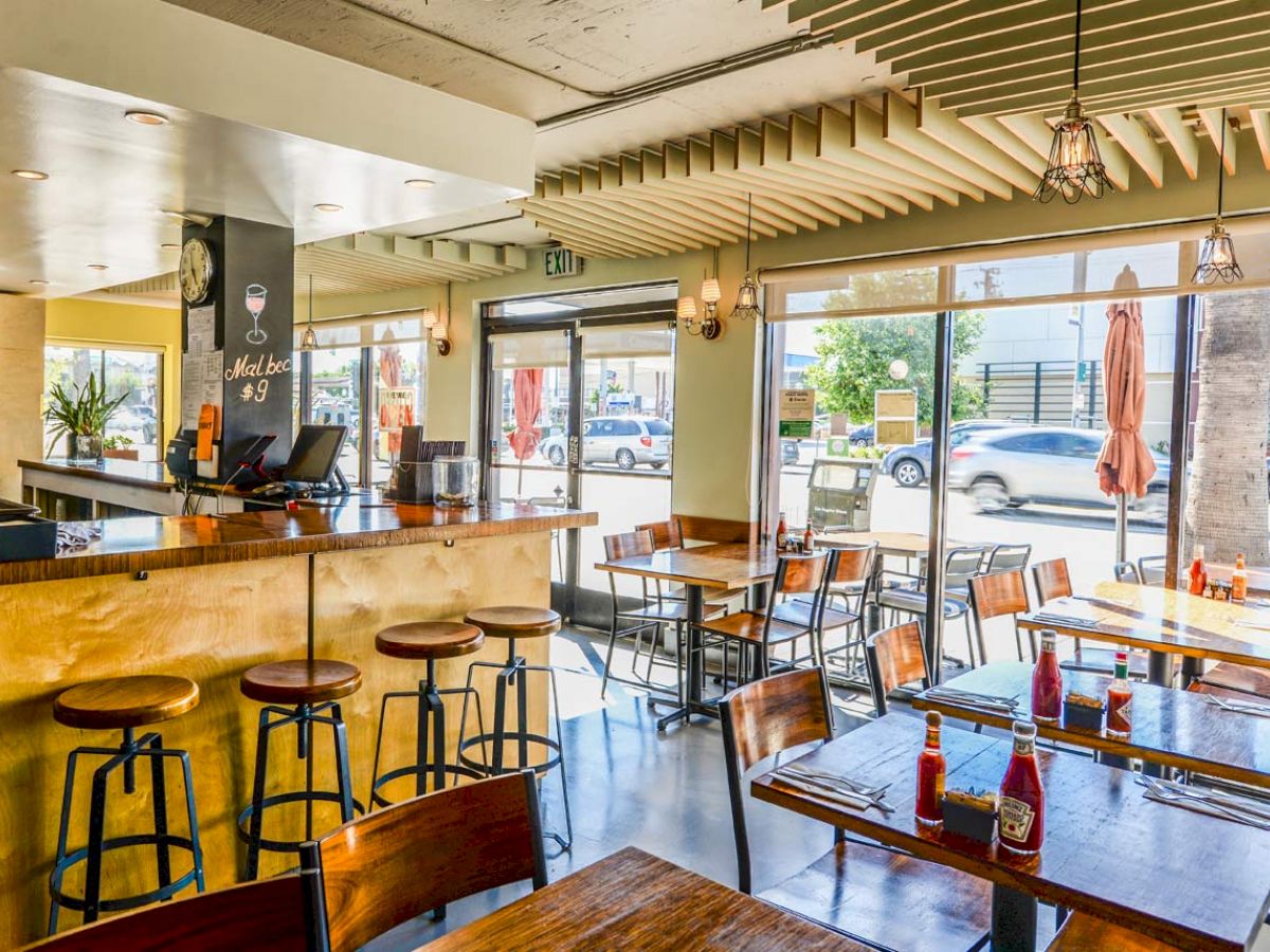 This is an image of a modern cafe with wooden tables, bar stools, condiment bottles on the tables, and large windows providing natural light.