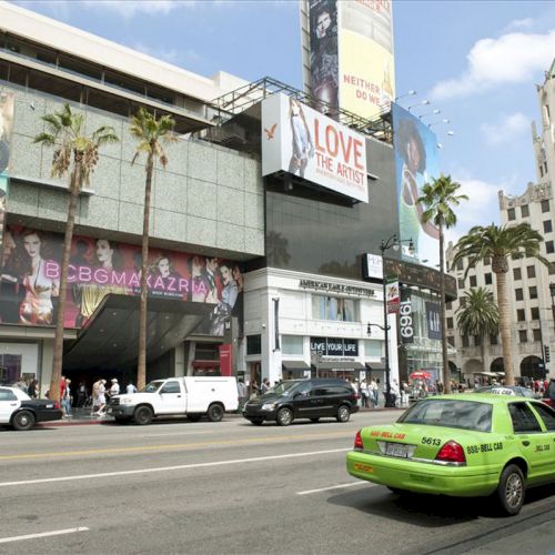 A busy street scene with cars, taxis, pedestrians, and various advertisements on buildings, possibly in a major city.