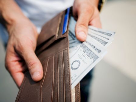 A person is holding an open brown leather wallet with several $100 bills partially visible inside.