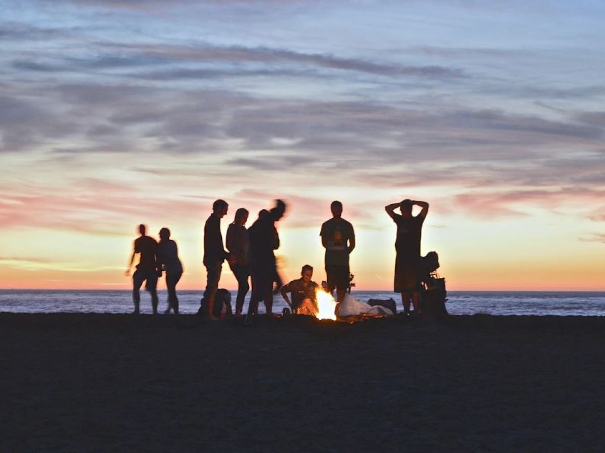 A group of people gathered around a bonfire on a beach at sunset, with the ocean and a colorful sky in the background.